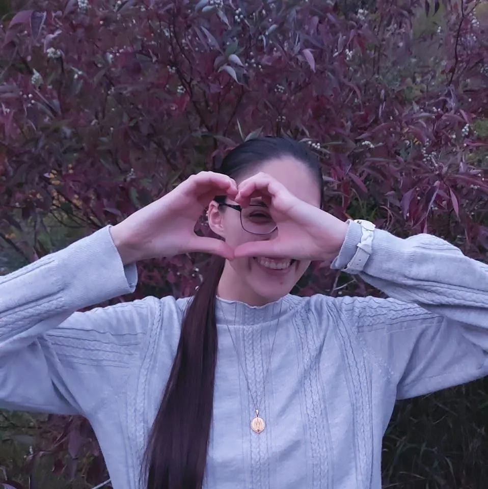 Rachel making a heart shape with her hands in front of deep red autumn leaves