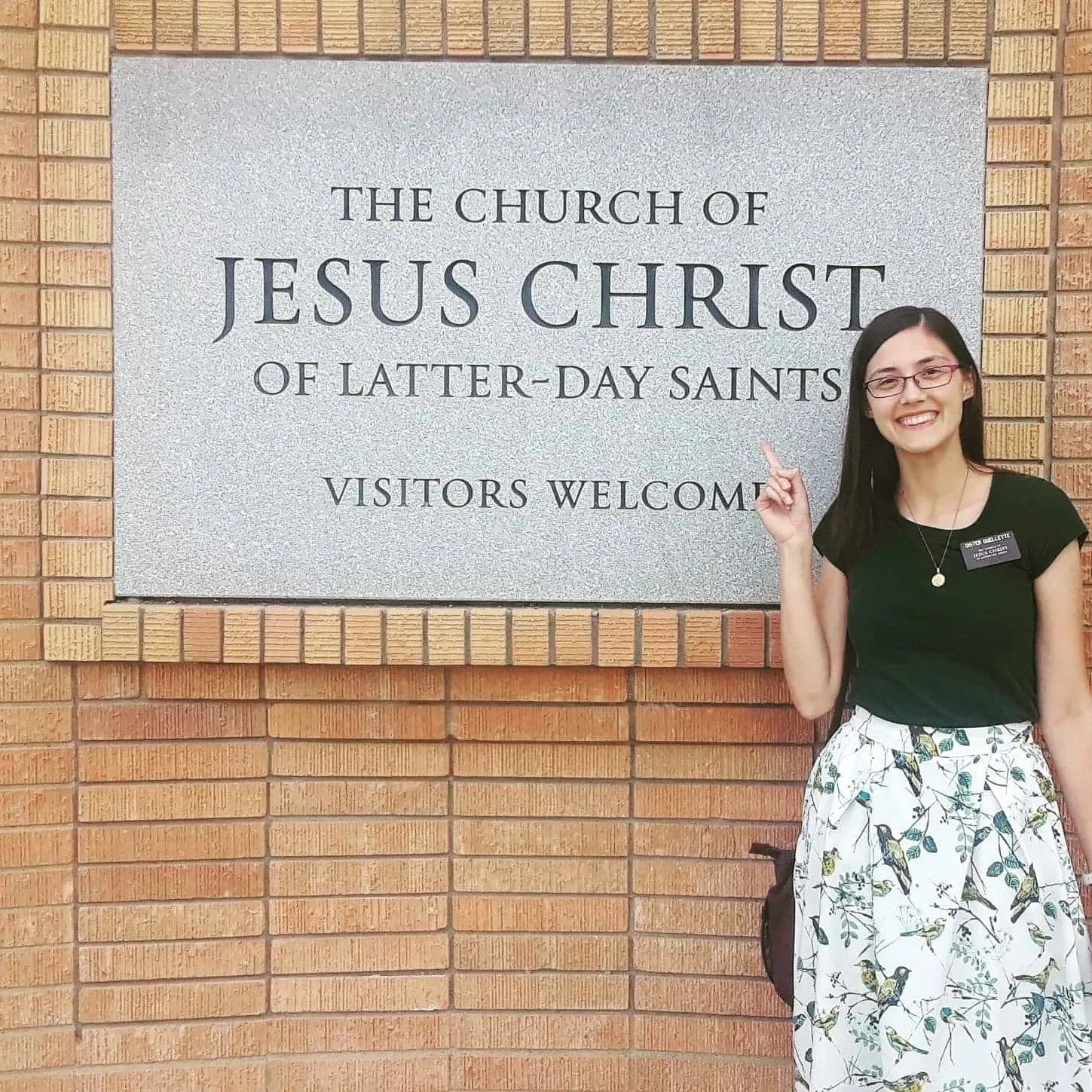 Rachel as a sister missionary pointing to a meetinghouse sign that reads "Visitors Welcome"