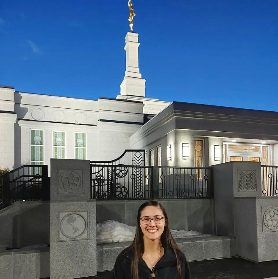 Rachel at the gate of a white temple topped with a gold angel Moroni against a deep blue sky