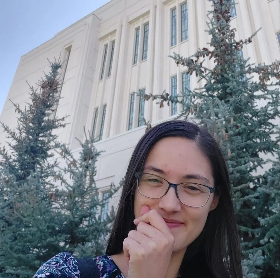 Rachel smiling with her hand at her chin beside a white temple and blue spruce trees